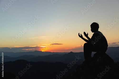 Silhouette of a young man praying to God on the mountain at sunset background. Woman raising his hands in worship. Christian Religion concept.