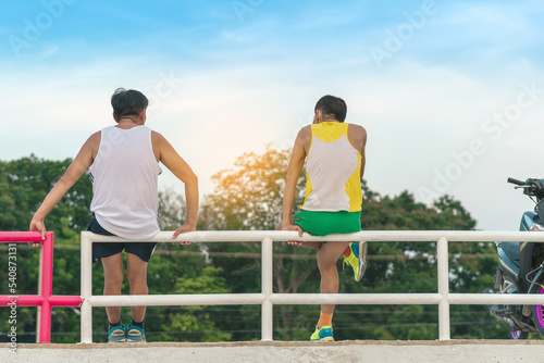 Back view of two sporty senior male friends in sportswear sitting together on railing fence in park talking about something. Time to rest. Social distance during the Corona Virus (COVID-19) pandemic.