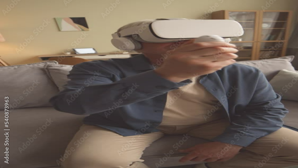 Vertical of excited gray haired man wearing vr headset using vr controller while playing vr games at home