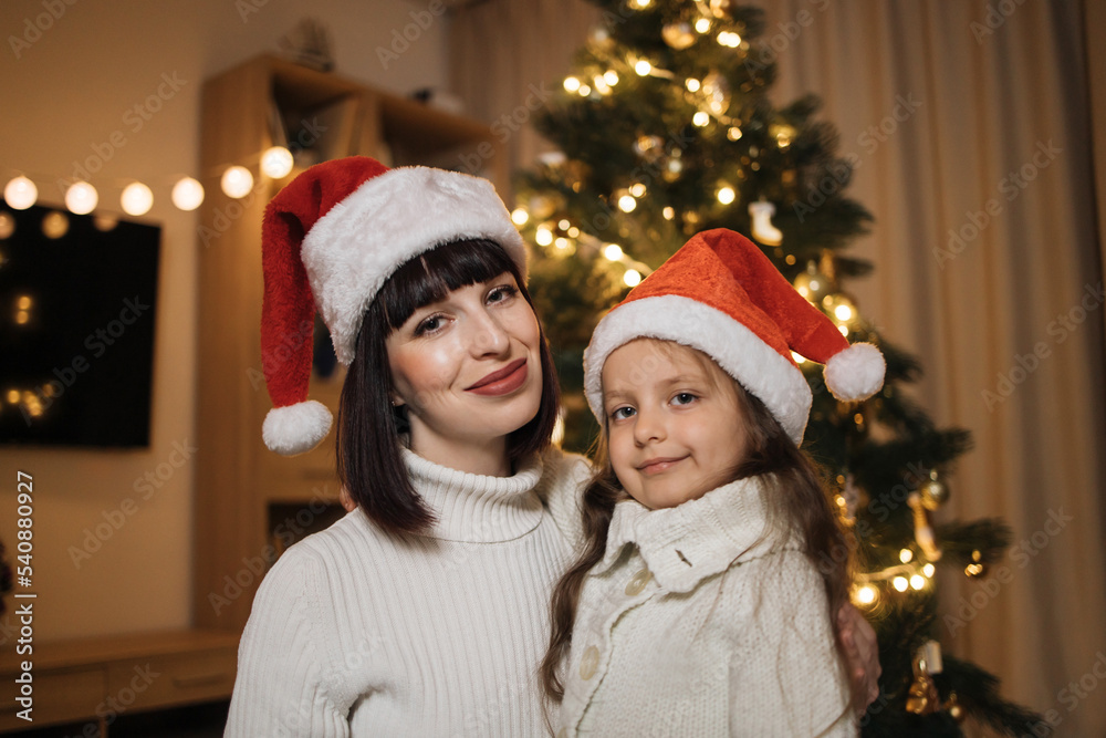Close up portrait of lovely family of two, beautiful young mother and her cute little daughter in warm knitted white sweaters in front of decorated Christmas tree. Close up portrait of lovely family of two, beautiful young mother and her cute little daughter in warm knitted white sweaters in front of decorated Christmas tree.