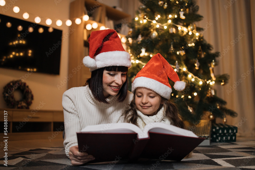 Pretty young mother in santa hat reading magical fairy tale book to her daughter lying on floor near Christmas tree, merry Christmas. Pretty young mother in santa hat reading magical fairy tale book to her daughter lying on floor near Christmas tree, merry Christmas.