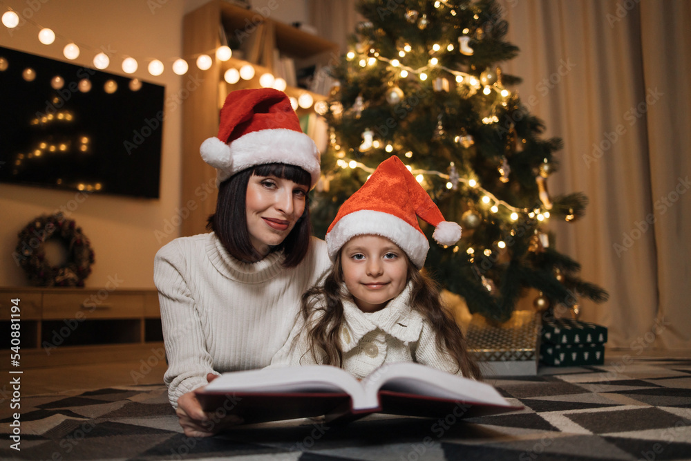 Close up view of lovely family of two, beautiful young mother and her cute little daughter in warm knitted white sweaters reading fairy tale book while lying in front of decorated Christmas tree. Close up view of lovely family of two, beautiful young mother and her cute little daughter in warm knitted white sweaters reading fairy tale book while lying in front of decorated Christmas tree.