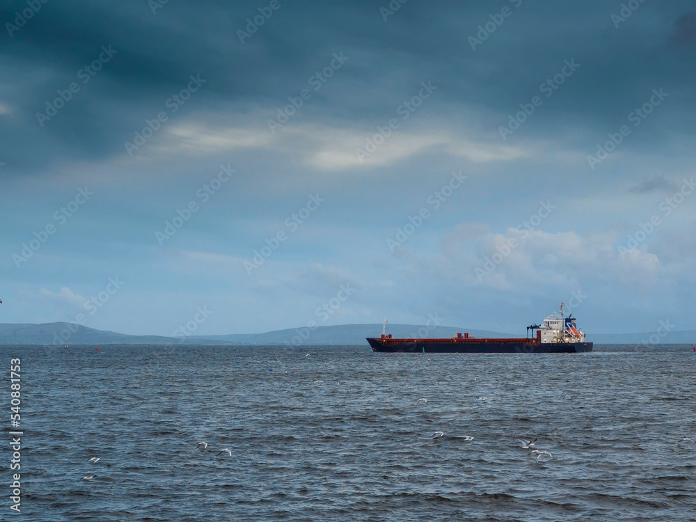 Cargo ship leaving port. Commercial carrier vessel delivering goods and products by water. Import and export concept. Transportation industry. Dramatic cloudy sky.