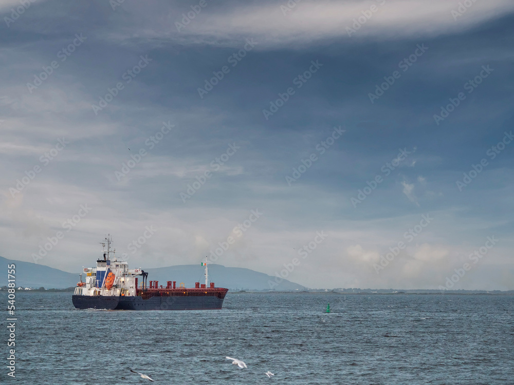 Cargo ship leaving port. Commercial carrier vessel delivering goods and products by water. Import and export concept. Transportation industry. Dramatic cloudy sky.