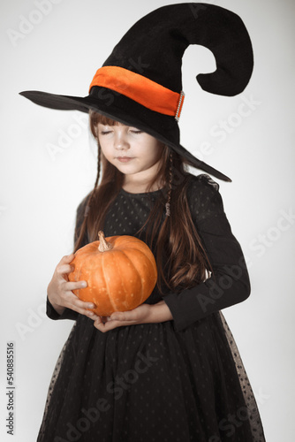 Halloween portrait of a small smiling witch in a black hat with pumpkin