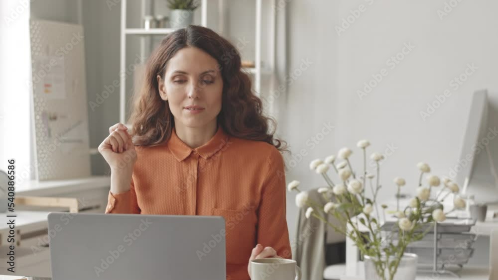 Young dreamy businesswoman enjoying her cup of coffee while working on laptop at bright cozy office Young dreamy businesswoman enjoying her cup of coffee while working on laptop at bright cozy office