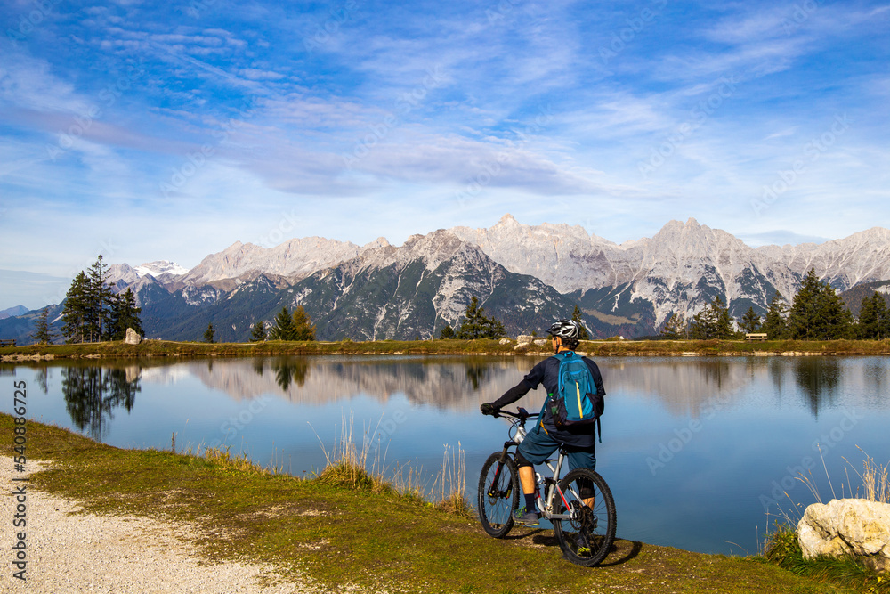 Mountainbiker macht eine Pause am Kaltwassersee in Seefeld/Tirol. Der schneebedeckte Gipfel am linken Bildrand ist die Zugspitze - obrazy, fototapety, plakaty 