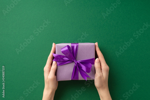New Year concept. First person top view photo of woman's hands giving violet giftbox with purple ribbon bow on isolated green background with blank space