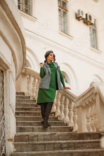 Outdoor fashion portrait of an elegant fashionable brunette model in a beret, green dress and a gray waistcoat posing at sunset in a european city in autumn.