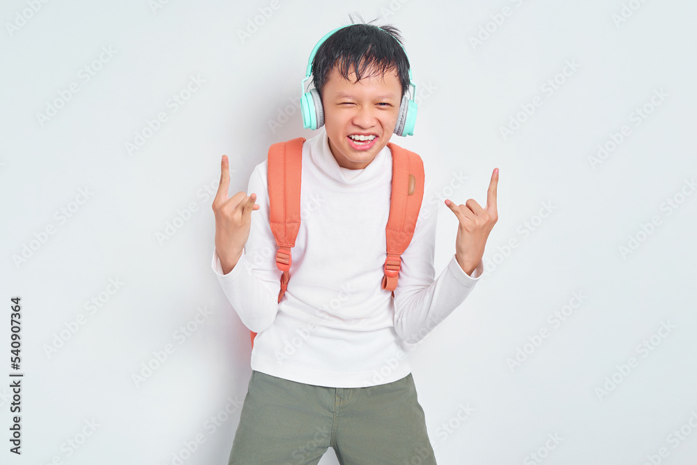 Rock and Roll. handsome young Asian student in Christmas hat and casual clothes with backpack, listening music rock with headphones and showing metal rock gesture isolated on white background Rock and Roll. handsome young Asian student in Christmas hat and casual clothes with backpack, listening music rock with headphones and showing metal rock gesture isolated on white background