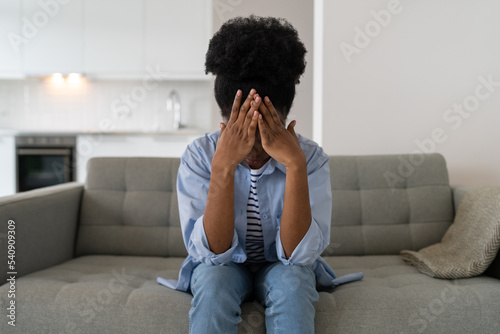 Confused African woman in casual shirt and jeans covers face with hands after quarrel with friends. Discouraged black female with problems in personal life sits on sofa in living room of apartment
