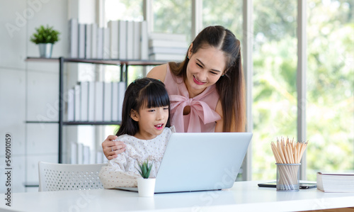 Millennial Asian happy family mother smiling helping supporting teaching little girl kid daughter studying learning doing online school homework via laptop notebook computer in living room at home