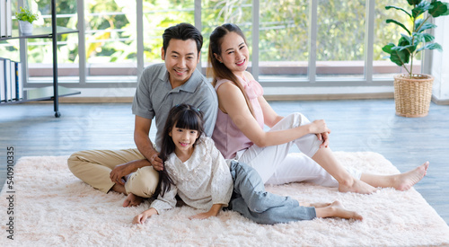 Millennial Asian happy family father mother and little girl daughter in casual outfit sitting laying lying down on fluffy carpet floor smiling posing together taking photograph in living room at home