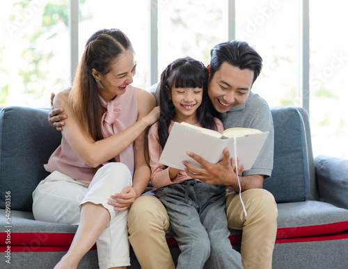 Millennial Asian happy family father mother sitting on cozy sofa couch smiling together teaching little girl kid daughter learning studying reading fiction novel textbook in living room at home