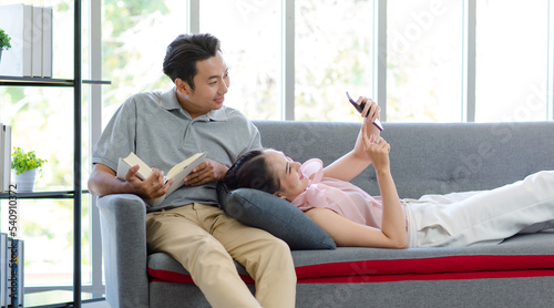 Millennial Asian happy family husband sitting smiling reading novel book on cozy sofa couch while lovely wife laying down on lap using smartphone video call talking with friend in living room at home