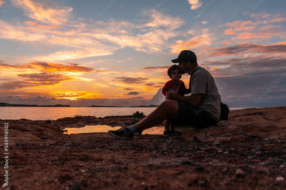 Sunset in Ibiza on vacation, young father giving his son a kiss by the sea, San Antonio Abad