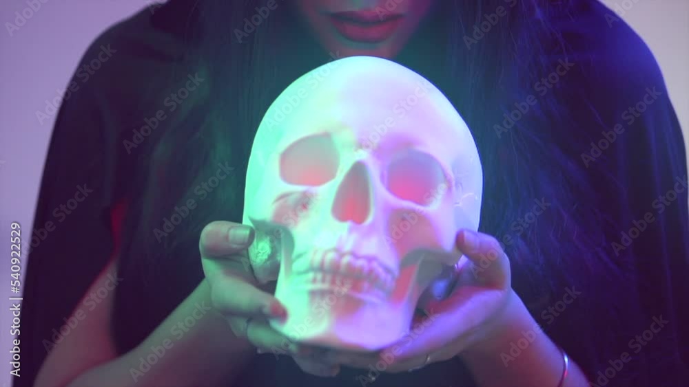 Halloween Witch woman holding human skull lantern in her hands. Beautiful young woman doing witchcraft at night. Darkness. Scary scene. Over spooky dark blue magic background Halloween Witch woman holding human skull lantern in her hands. Beautiful young woman doing witchcraft at night. Darkness. Scary scene. Over spooky dark blue magic background