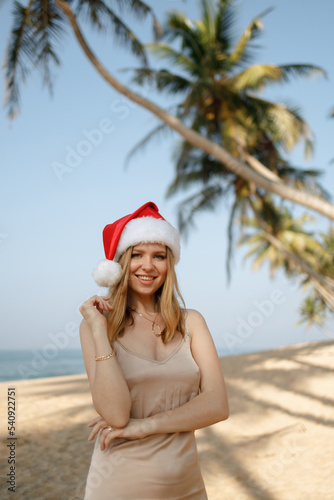 Beautiful girl in a Santa Claus hat on the beach with palm trees. Christmas on the tropical coast.