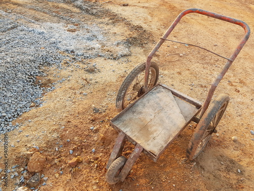 Wheelbarrow on construction area, Metal trolley with three wheels, Rusty old cart on the background of piles of stones and sand.