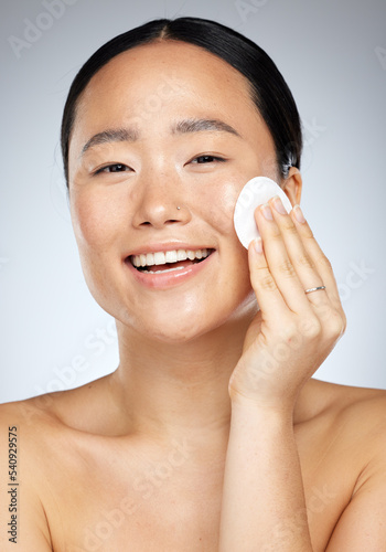 Skincare, beauty and woman with cotton pad for facial cleaning against a grey studio background. Happy, smile and portrait of a young Asian model with cotton for face wellness, clean body and makeup