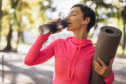 Fit woman srinking water in park and holding yoga mat photo