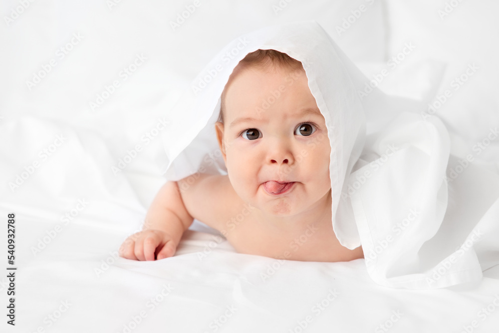 Funny baby lying on bed peeking under white sheet sticking tongue out Funny baby lying on bed peeking under white sheet sticking tongue out