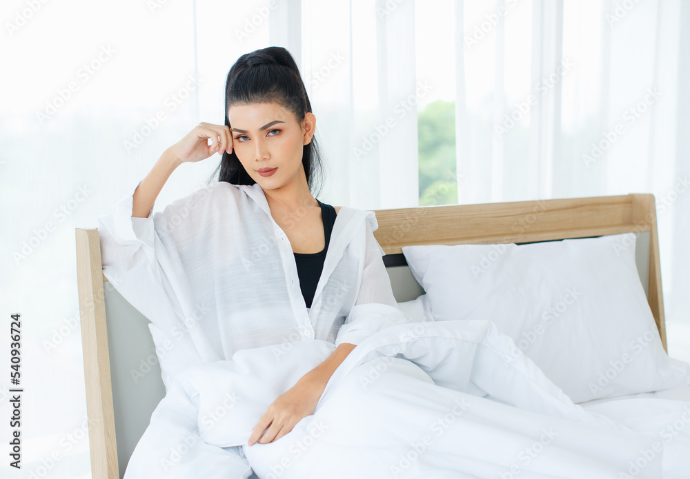Portrait shot of Millennial Asian sexy female in casual comfort tanktop and shirt sitting under white thick warm blanket posing look at camera smiling after waking up in bedroom in morning at home Portrait shot of Millennial Asian sexy female in casual comfort tanktop and shirt sitting under white thick warm blanket posing look at camera smiling after waking up in bedroom in morning at home