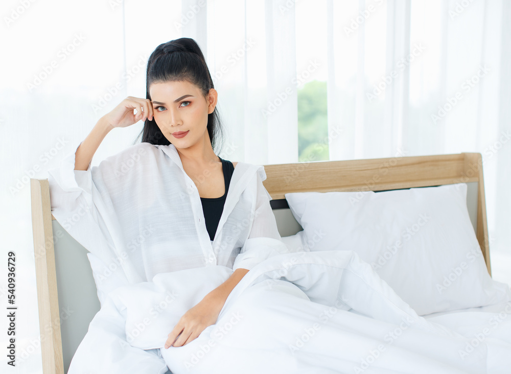 Portrait shot of Millennial Asian sexy female in casual comfort tanktop and shirt sitting under white thick warm blanket posing look at camera smiling after waking up in bedroom in morning at home Portrait shot of Millennial Asian sexy female in casual comfort tanktop and shirt sitting under white thick warm blanket posing look at camera smiling after waking up in bedroom in morning at home
