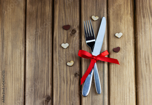 Romantic dinner concept. Top view of silver fork and knife decorated red ribbon with bow on wooden background