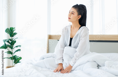 Millennial Asian sexy thoughtful female in casual comfort pajamas sitting under white thick warm blanket starting day thinking looking outside alone after waking up in bedroom in morning at home