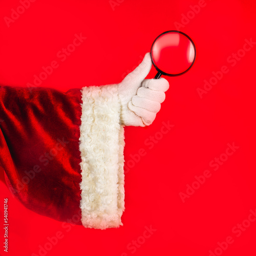 Santa's hand in a traditional red suit and white glove holds a magnifying glass, a symbol of research, search, observation, control, intention, goals of the year.