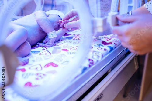 Unrecognizable mother hands caressing and touching her new born baby in intensive care unit in a medical incubator under ultraviolet lamp.