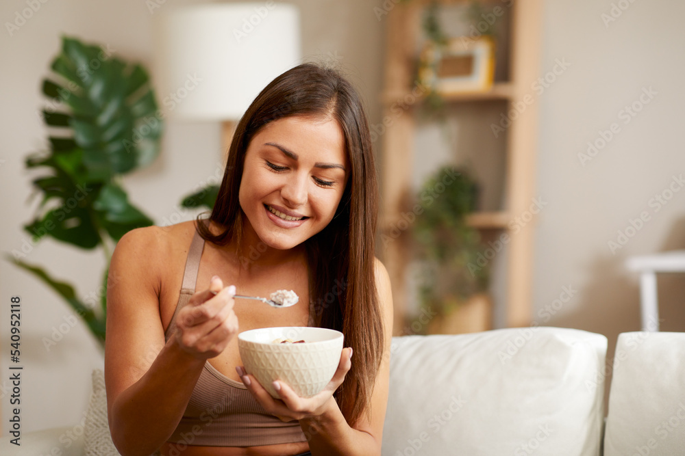 Beautiful young woman is sitting on the sofa in the living room and eating oatmeal Beautiful young woman is sitting on the sofa in the living room and eating oatmeal