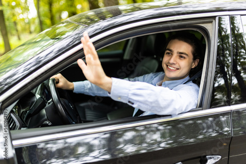 Portrait of young handsome brunette man driving car and greeting somebody with hand.