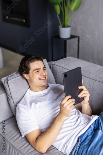 Young man with beard lying on the sofa and holding tablet in hands. Man indoor with laptop tablet. Hipster man relaxing and looking to the tablet. Young man with tablet.