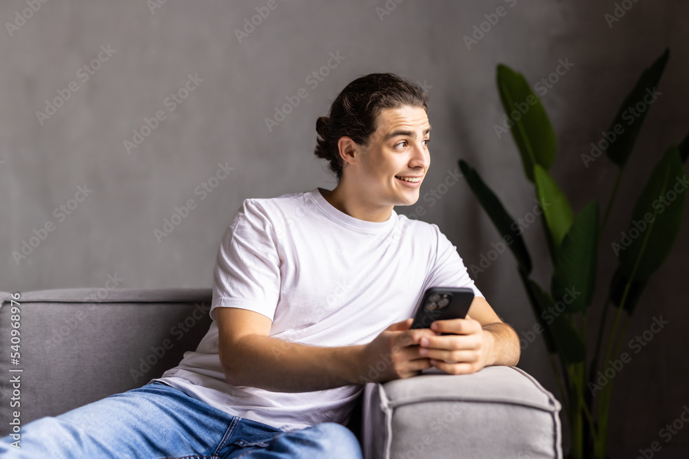 Portrait of an attractive smiling young man wearing casual clothes sitting on a couch at the living room, using mobile phone Portrait of an attractive smiling young man wearing casual clothes sitting on a couch at the living room, using mobile phone
