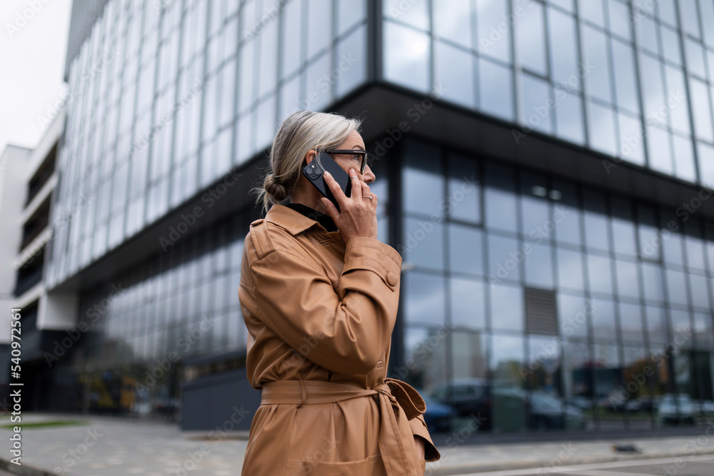 Fototapeta premium Business senior adult woman in an autumn coat on the background of the glass facade of the office center speaks on a mobile phone, 50 year old European woman, business strategy concept
