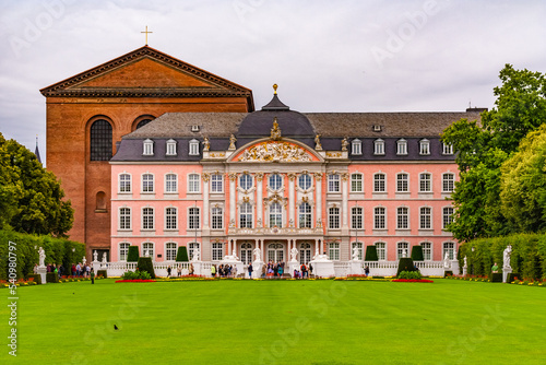 Famous view of the Electoral Palace in Trier, Germany with the basilica behind it. The lawn and flower beds in front of the main entrance to the south wing are bordered by hedges and trees.