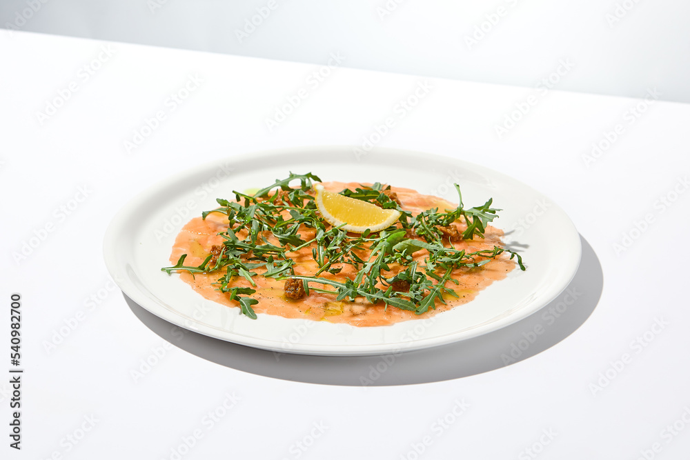 Salmon carpaccio with arugula on white background in summer day. Fish appetizer - salmon carpaccio in fine dining with shadows. Elegant carpaccio of raw salmon and sauce. Italian food. Aesthetic menu. Salmon carpaccio with arugula on white background in summer day. Fish appetizer - salmon carpaccio in fine dining with shadows. Elegant carpaccio of raw salmon and sauce. Italian food. Aesthetic menu.