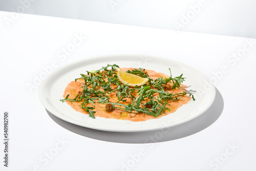 Salmon carpaccio with arugula on white background in summer day. Fish appetizer - salmon carpaccio in fine dining with shadows. Elegant carpaccio of raw salmon and sauce. Italian food. Aesthetic menu.