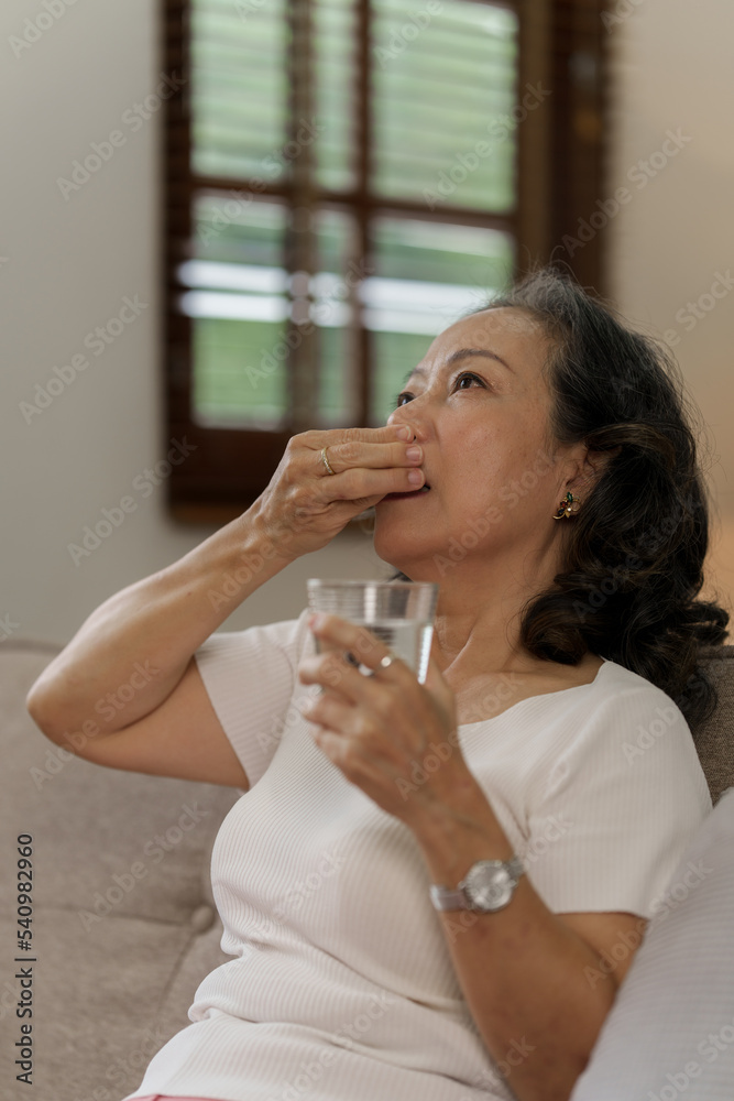 Elderly Southeast Asian woman holding heart disease pills sitting on the sofa alone, elderly health concept Elderly Southeast Asian woman holding heart disease pills sitting on the sofa alone, elderly health concept