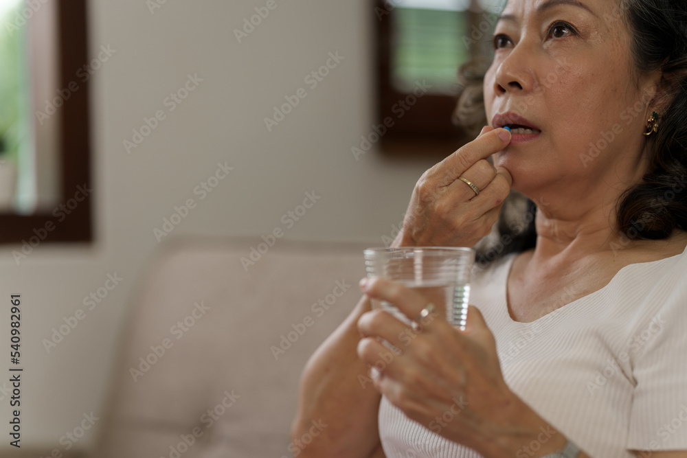 Elderly Southeast Asian woman holding heart disease pills sitting on the sofa alone, elderly health concept Elderly Southeast Asian woman holding heart disease pills sitting on the sofa alone, elderly health concept