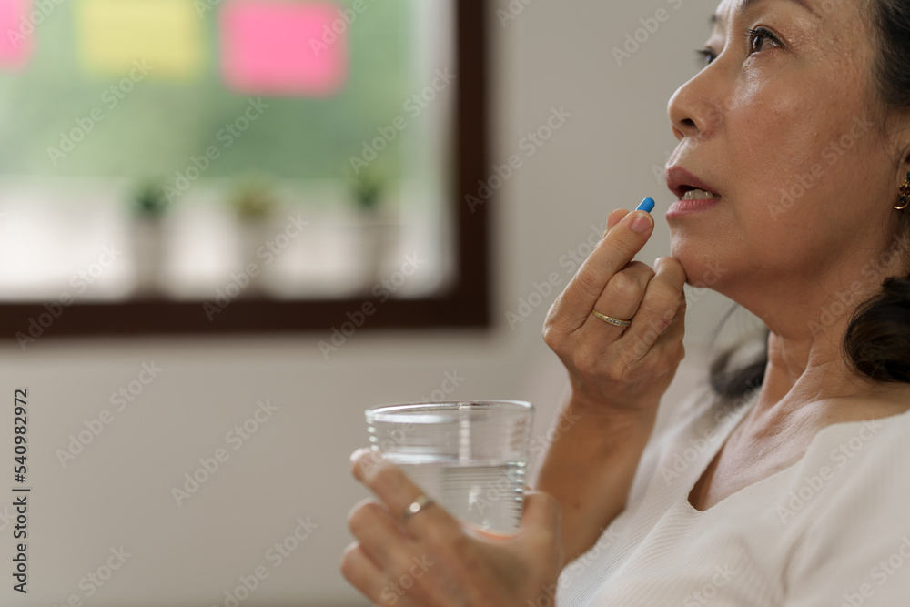 Elderly Southeast Asian woman holding heart disease pills sitting on the sofa alone, elderly health concept