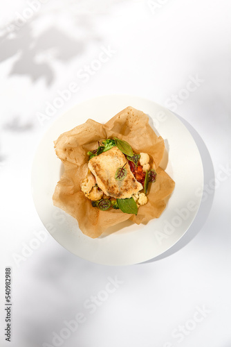 Baked cod fillet with vegetables in parchment. Roasted cod steak with cauliflower. Healthy food in summer menu. White fish roasted in parchment. Fish dish in white background with shadows.