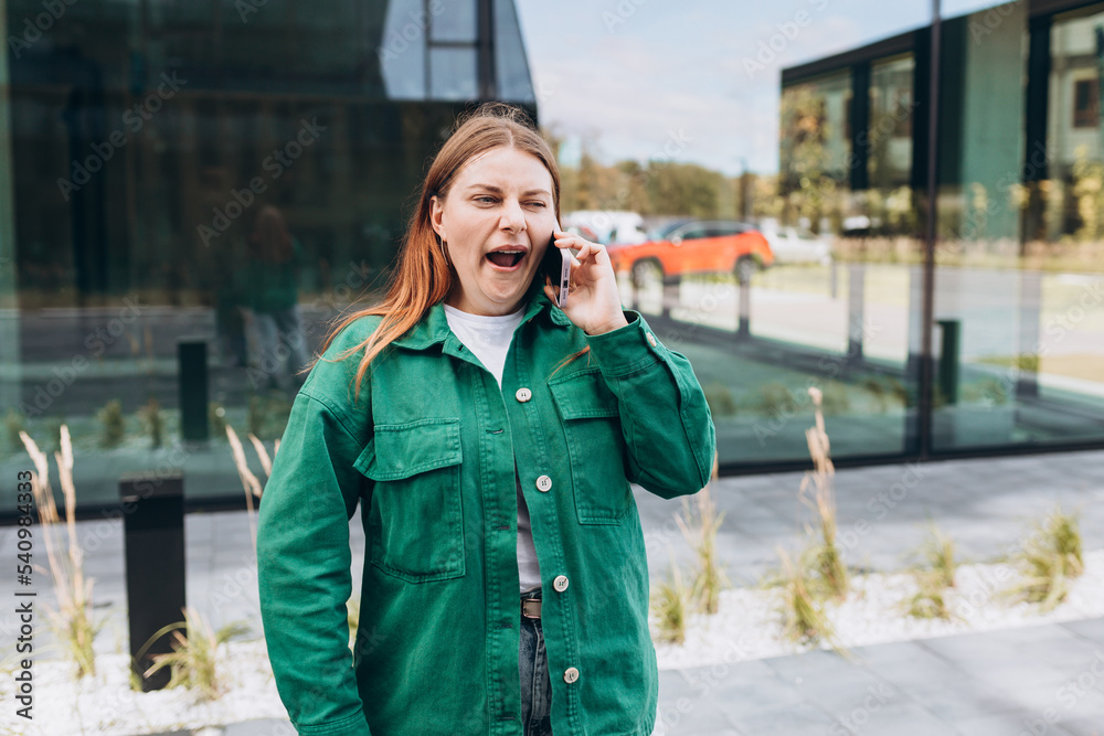 Fototapeta premium Beautiful Bored redhead woman talking with her smartphone. Woman yawning and using smartphone on city street.