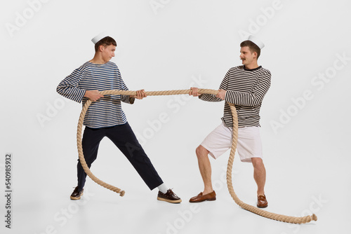 Portrait of two young boys, sailor, seamen in striped shirts pulling rope isolated over white background. Competition