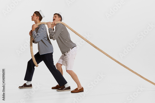 Portrait of two young boys, sailor, seamen in striped shirts pulling rope isolated over white background