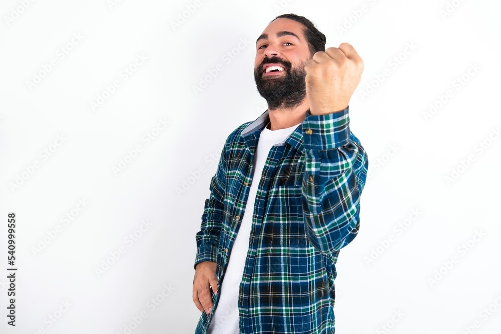 Fototapeta premium Overjoyed young bearded hispanic man wearing plaid shirt over white background glad to receive good news, clenching fist and making winning gesture.