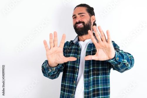 Dissatisfied young bearded hispanic man wearing plaid shirt over white background frowns face, has disgusting expression, shows tongue, expresses non compliance, irritated with somebody.