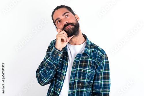 Face expressions and emotions. Thoughtful young bearded hispanic man wearing plaid shirt over white background holding hand under his head, having doubtful look.